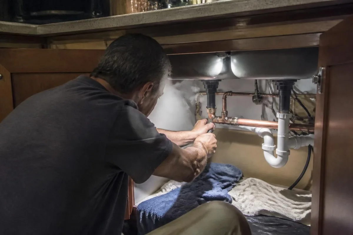 Plumber repairing a split copper pipe under a kitchen sink at night, towels on the floor, emergency plumber in Bailey, NC